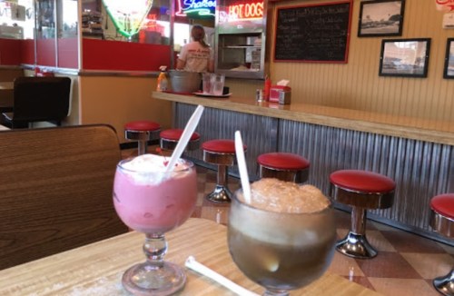 Two dessert glasses with pink and brown drinks sit on a table in a retro diner, with red stools and a neon sign in the background.