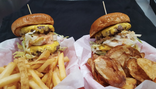 Two stacked cheeseburgers with lettuce and sauce, served with fries and potato chips in a casual dining setting.