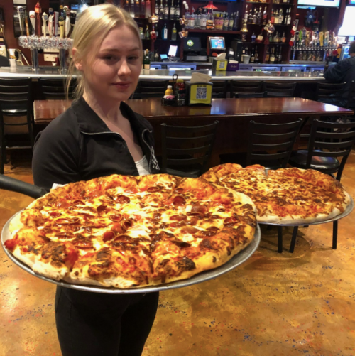 A server holds two large pizzas on a tray in a restaurant with a bar in the background.