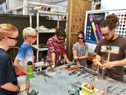 A group of four kids and an adult work together on glassblowing, wearing safety glasses in a workshop.