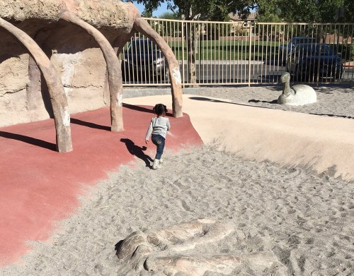 A child walks on sandy ground near a playground structure resembling rocks and dinosaur bones.