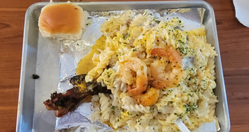 A tray with creamy pasta topped with shrimp, a piece of bread roll, and a side of grilled meat.