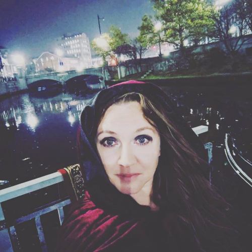 A woman in a dark hooded cloak stands by a canal at night, with city lights reflecting on the water.