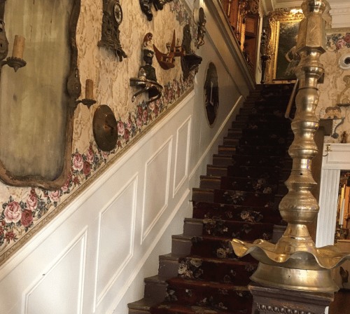 A vintage staircase with floral wallpaper and antique decor, leading upward in a warmly lit space.