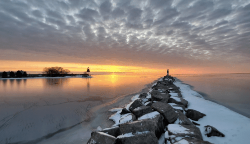 A serene winter sunset over a frozen lake, with a rocky pier leading to a distant lighthouse and a person standing at the end.