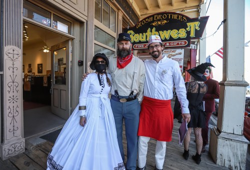 Three people in costumes pose outside a store: a woman in a white dress, a man in a Confederate uniform, and another in a red apron.