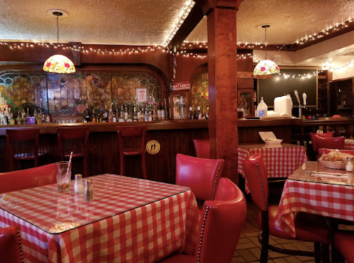 Cozy restaurant interior with checkered tablecloths, red chairs, and a bar adorned with lights and stained glass.