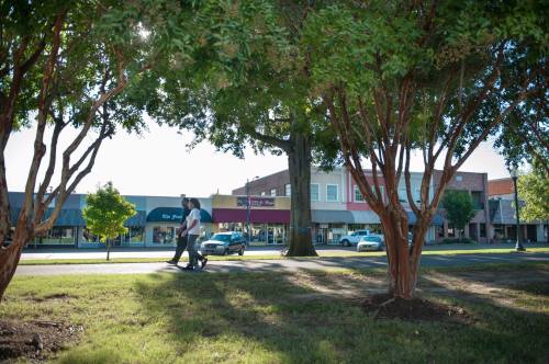 Two people walk along a tree-lined street with shops in the background on a sunny day.