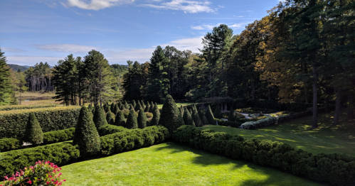 Lush green garden with neatly trimmed hedges and trees, under a clear blue sky, surrounded by a scenic landscape.