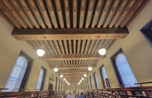 A library interior featuring a decorative ceiling with wooden beams and hanging lights, and bookshelves in the background.