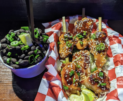 A plate of glazed chicken skewers garnished with sesame seeds and green onions, alongside a bowl of black beans.