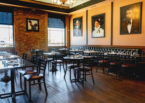A cozy restaurant interior with wooden floors, empty tables, and vintage portraits on the walls.