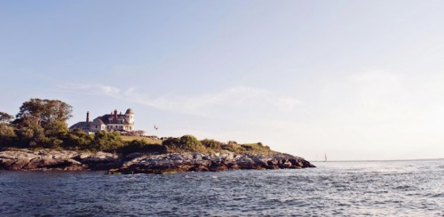 A coastal view of a large house on a rocky shore, with calm waters and a clear sky in the background.