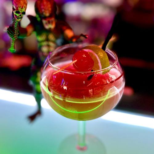 A colorful cocktail in a large glass, garnished with a cherry and lime, with a toy figure in the background.