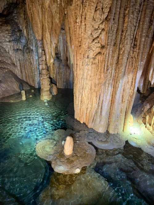 Stalactites and stalagmites in a dimly lit cave, with clear water reflecting the formations.