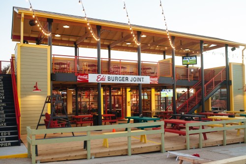 A two-story burger joint with outdoor seating, colorful tables, and string lights, featuring a large sign that reads "Ed's Burger Joint."
