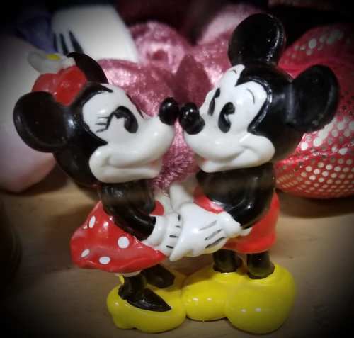 Two cartoon mice figures, Mickey and Minnie, holding hands and gazing at each other, both wearing their iconic outfits.