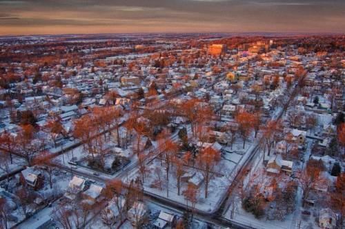 Aerial view of a snowy suburban landscape at sunset, with houses and trees reflecting warm colors in the light.