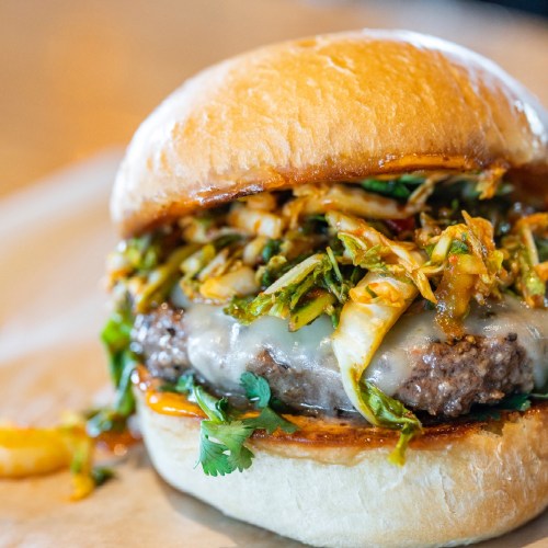 A close-up of a juicy burger topped with shredded vegetables and cilantro, served on a soft bun.