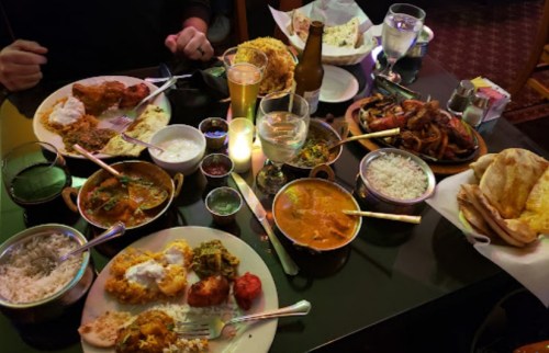 A table filled with various Indian dishes, including curries, rice, naan, and drinks, in a cozy restaurant setting.