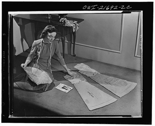 A woman examines fabric patterns laid out on the floor, with a magazine and sewing materials nearby.