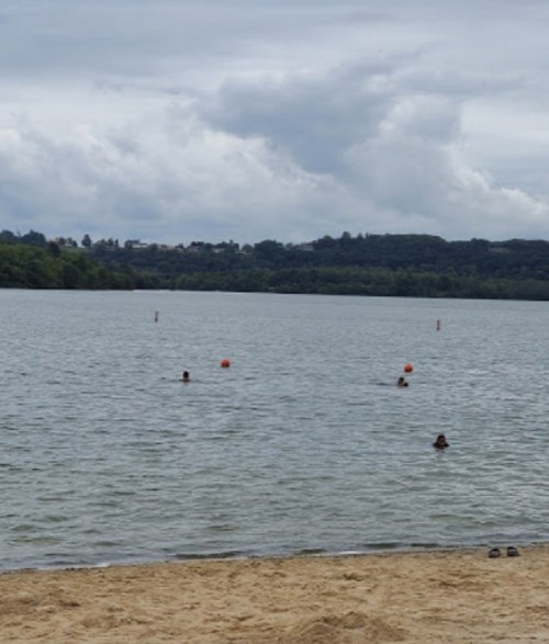 A calm lake with a sandy shore, people swimming, and overcast skies in the background.
