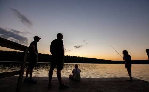 Silhouettes of four people on a dock at sunset, with one person fishing by the water's edge.