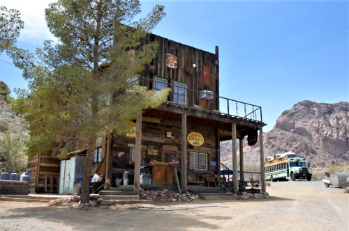 A rustic wooden building with a porch, surrounded by trees and mountains, in a desert landscape.