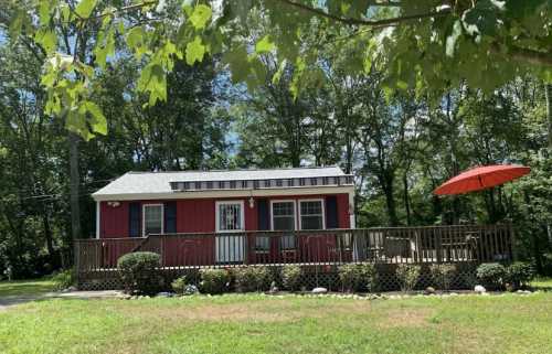 A small red cottage with a porch, surrounded by greenery and featuring an orange umbrella on the deck.