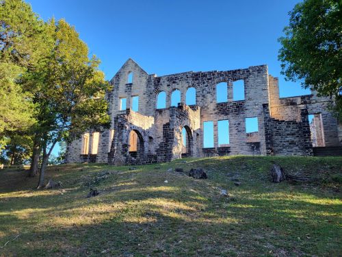 Ruins of a stone building with arched windows, surrounded by trees and grass under a clear blue sky.