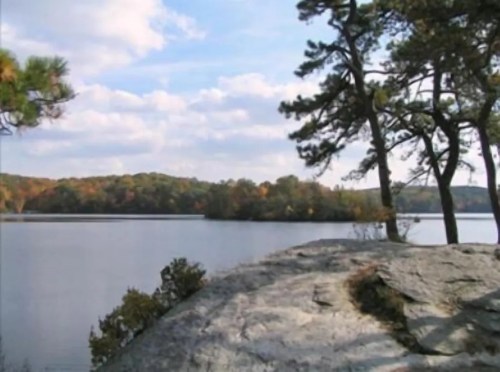 A serene lake surrounded by trees and rocky terrain under a partly cloudy sky.