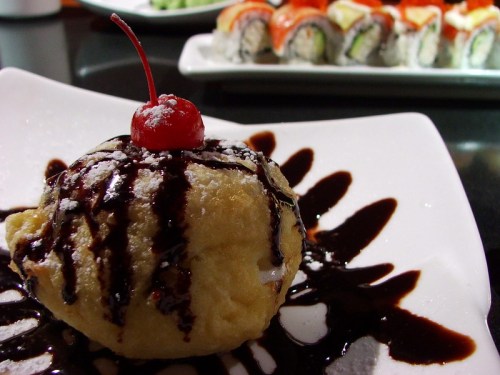 A dessert ball drizzled with chocolate sauce and topped with a cherry, served on a white plate with sushi in the background.