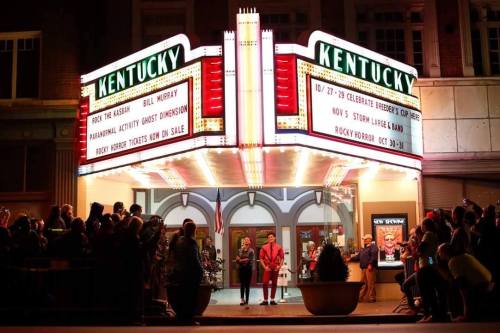 A vibrant theater marquee reads "Kentucky," with a crowd gathered outside for an event. Bright lights illuminate the scene.