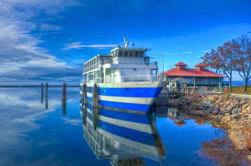 A blue and white ferry docked at a calm harbor, reflecting in the water under a clear blue sky.