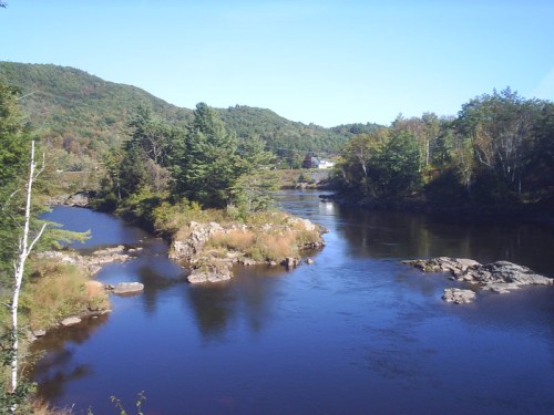 A serene river flows through a lush landscape, surrounded by trees and rocky outcrops under a clear blue sky.