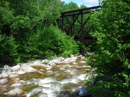 A rustic metal bridge spans a rocky river, surrounded by lush green trees and a clear blue sky.