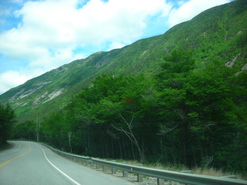 A winding road alongside lush green mountains under a partly cloudy sky.
