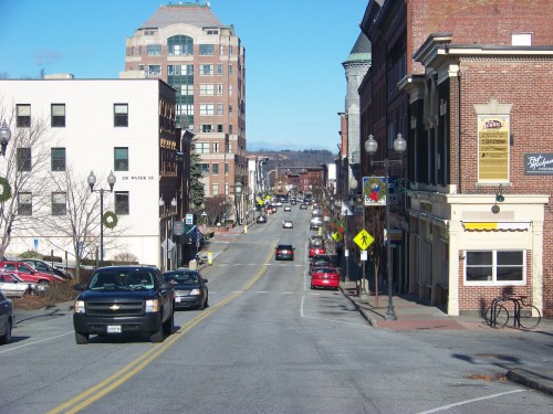 A city street lined with buildings, cars parked along the sides, and clear blue skies overhead.