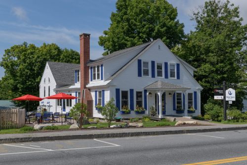 A charming white house with blue shutters, surrounded by greenery, featuring a patio with red umbrellas.