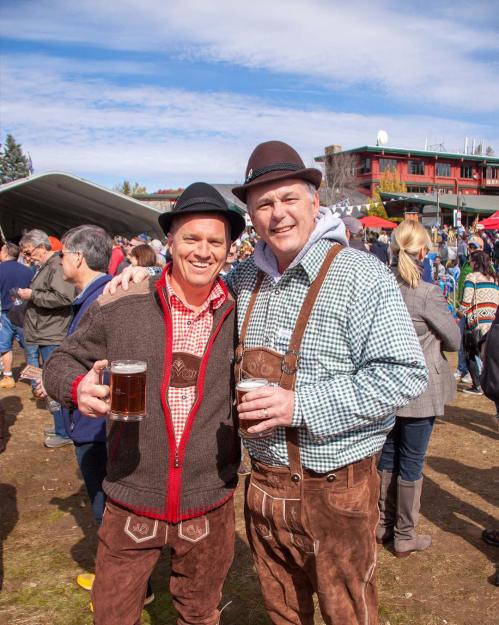 Two men in traditional attire hold beers at a lively outdoor festival, with a crowd and tents in the background.