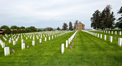 A serene cemetery with white headstones and small American flags, set against a backdrop of green grass and trees.