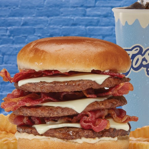 A towering burger with two beef patties, melted cheese, and crispy bacon, served with fries and a drink in the background.