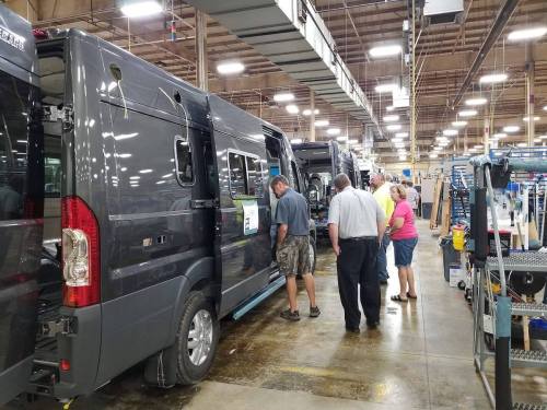 A group of people inspecting gray vans in a spacious manufacturing facility.