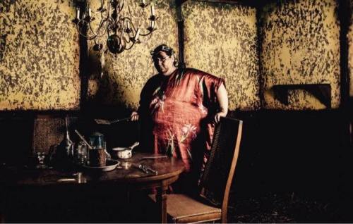A person in a red floral robe stands in a dimly lit, worn room with a table set for a meal.