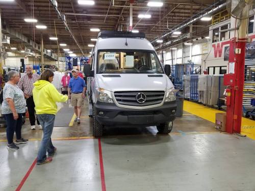 A group of people observes a silver Mercedes van in a factory setting, with equipment and workstations in the background.