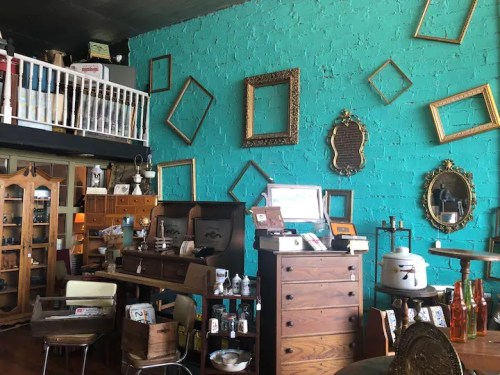 A vibrant turquoise wall adorned with empty frames, surrounded by vintage furniture and decor in a cozy shop.