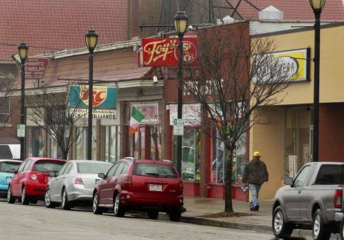 A street scene featuring shops, parked cars, and a person walking, with storefronts and lampposts lining the sidewalk.
