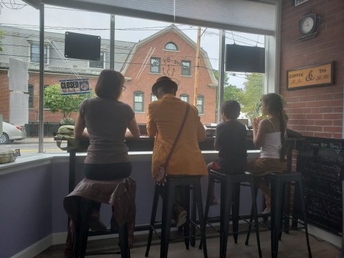 Four people sit at a counter in a café, facing a window with a view of a street and a closed sign.