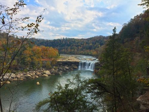 A serene landscape featuring a waterfall cascading into a river, surrounded by autumn foliage and rolling hills.