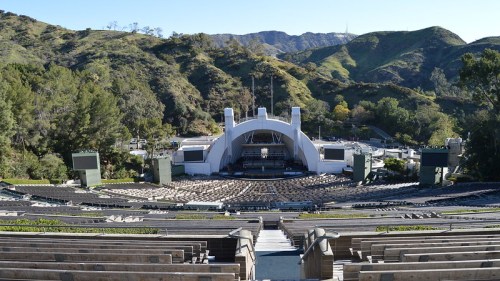 A wide view of an outdoor amphitheater surrounded by green hills, with empty seating and a stage set up for a performance.